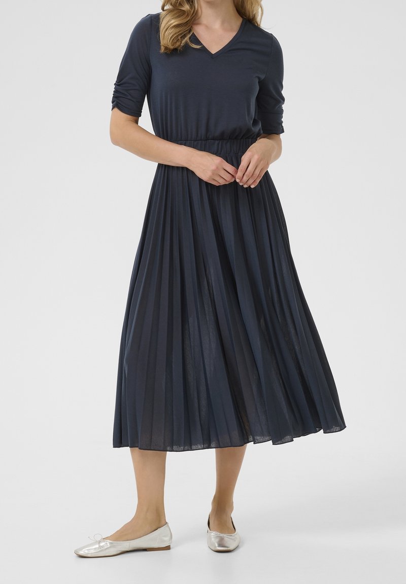 Robe plissée marine avec un col en V et des manches trois-quarts, accompagnée de ballerines argentées. Texture lisse et silhouette fluide.