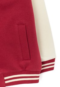 Detalle en primer plano de la manga y el bolsillo de una chaqueta varsity en color burdeos y blanco, con puños acanalados que presentan detalles de rayas blancas.
