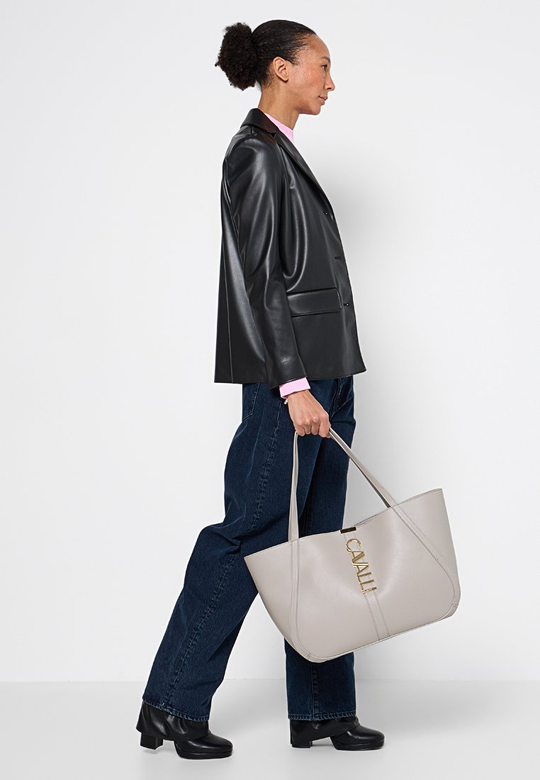 Frau in schwarzer Lederjacke und dunkler Jeans, die eine hellgraue Tote-Bag mit goldenen Buchstaben und schwarzen Schuhen hält, geht nach rechts.
