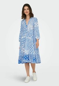 Abito blu a fantasia con scollo a V, maniche lunghe e gonna a strati. Realizzato in un tessuto leggero, presenta vari design blu e accenti bianchi.