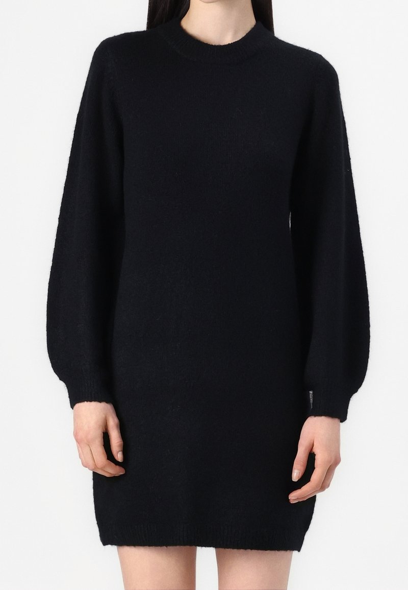 Femme portant une robe-pull en maille noire à manches longues avec un col rond sur un fond uni.
