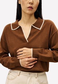 Pull en mélange de laine brun avec un col en V, un col blanc et des poignets. Texture douce avec des manches longues et une coupe décontractée. Fond de couleur neutre.