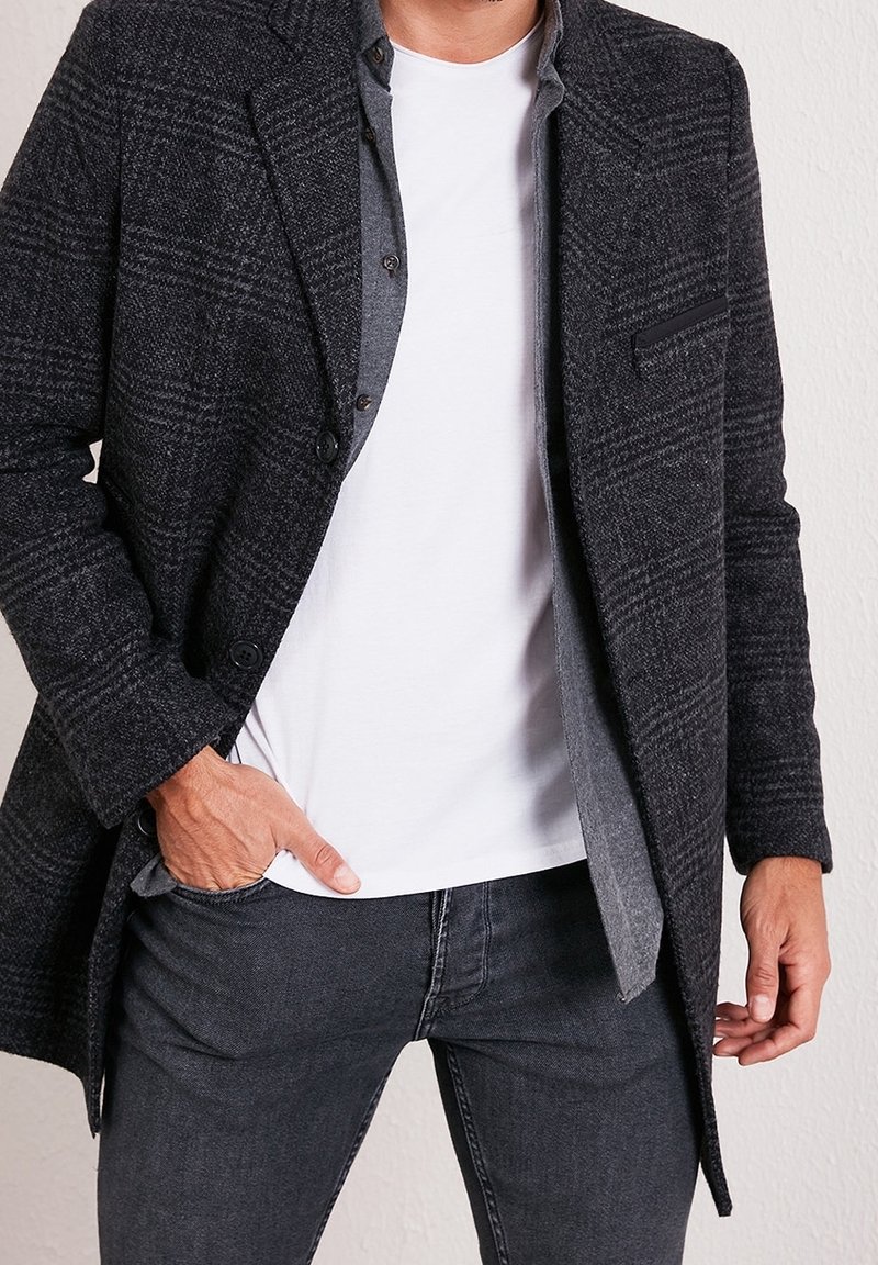 Homme avec la main dans la poche portant un manteau noir à carreaux, une chemise grise en dessous, un t-shirt blanc et un jean foncé devant un mur blanc.