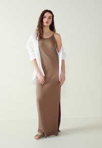 Rochie maxi maro cu bretele subțiri și o fanta pe o parte, asociată cu o cămașă albă deschisă și sandale maro deschis. Țesătură fină și design ajustat.