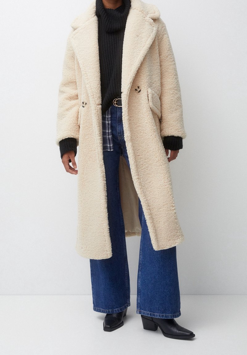 Personne portant un long manteau teddy crème, un pull à col roulé noir, un jean large bleu et des bottes à talons noires sur un fond blanc uni.