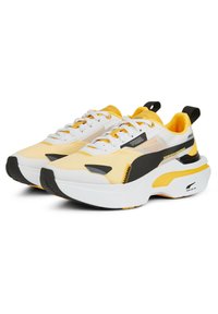 Sneakers bianchi, gialli e neri, realizzati con materiali in mesh e sintetici, suole ammortizzate, spesse suole bianche e lacci prominenti.