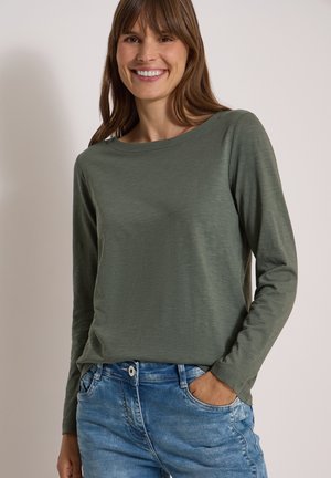 Femme souriante portant un haut à manches longues vert olive et un jean bleu avec une main dans la poche, sur un fond clair uni.