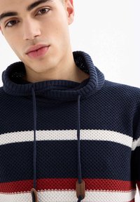 Joven vistiendo una sudadera con capucha de punto texturizado azul marino con rayas horizontales blancas y rojas, inclinado ligeramente contra un fondo liso.