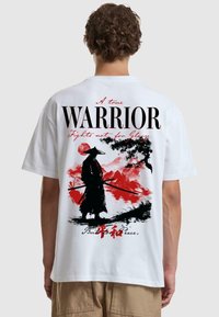 T-shirt bianco in cotone con una grande grafica nera e rossa di un samurai e il testo "Un vero GUERRIERO" e "Combatte non per la Gloria."