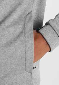 Grå sweatshirt med ribbad struktur, detalj med sidoficka och en figursydd mudd. Tyget verkar mjukt och andningsbart.