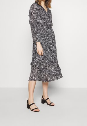 Frau trägt ein schwarzes Midikleid mit weißen Polka-Dots und Rüschen-Details, kombiniert mit schwarzen Sandalen mit Blockabsatz, vor einem schlichten Hintergrund.