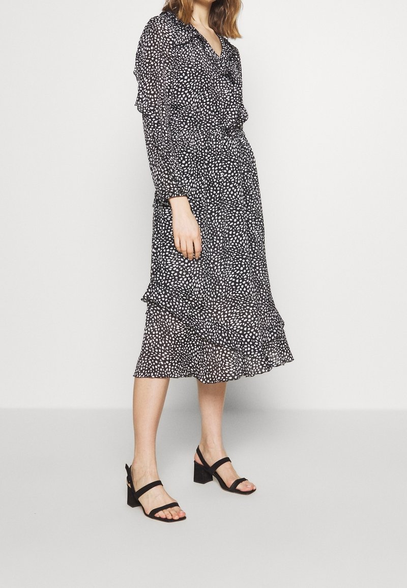 Femme portant une robe midi noire à pois blancs avec des détails à volants, associée à des sandales à talons blocs noires, debout devant un fond uni.