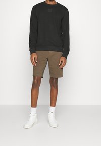 Czarny sweatshirt z okrągłym dekoltem z logo z przodu, w połączeniu z brązowymi szortami. Model nosi białe sneakersy za kostkę i skarpetki, stojąc na jednolitym tle.
