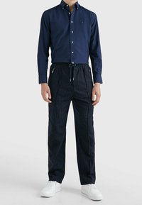 Hombre vistiendo una camisa de botones azul marino, pantalones oscuros de rayas con cordón y bolsillos con cremallera, y zapatillas blancas contra un fondo claro y liso.