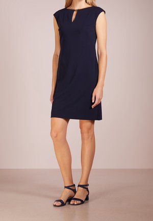 Kobieta ubrana w granatową sukienkę bez rękawów do kolan z małym otworem z przodu oraz granatowe sandały na słupku z paskiem wokół kostki.