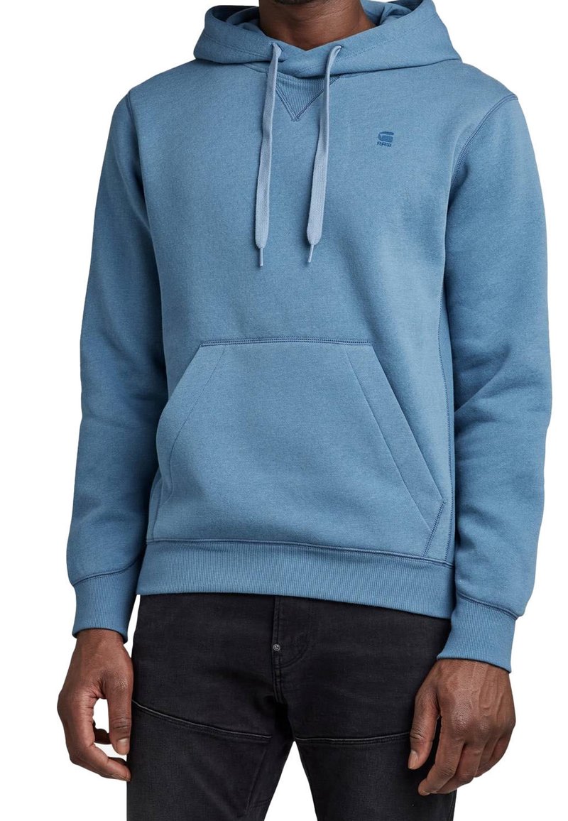 Homme portant un sweat à capuche bleu clair avec cordons et une poche kangourou devant, associé à un pantalon noir.