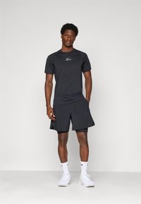 Mann står iført svart Nike Pro kortermet skjorte, svarte lagdelte shorts, hvite Nike-sokker og hvite joggesko mot en ensfarget bakgrunn.