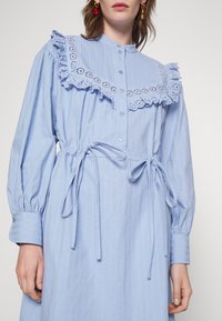 Ljusblå bomullsklänning med ribbad textur, utrustad med rufsat spetskrage, knappstängning fram och knytmidja. Långa ärmar med manschettdetaljer.