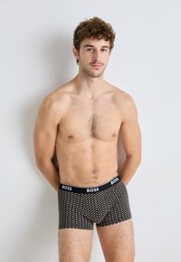 Schwarze Boxershorts mit weißen Punkten, elastischer Bund mit "BOSS"-Logo. Modell steht mit Händen hinter dem Rücken in einem neutralen Setting.
