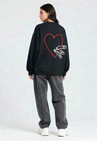 Črn oversized pulover s vezenim rdečim srcem in belimi varnostnimi sponkami na hrbtni strani, v kombinaciji s sivo ohlapnimi kavbojkami in belimi supergami.
