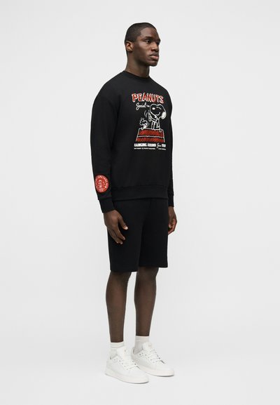 Άνδρας που στέκεται, φορώντας μαύρο φούτερ Peanuts με τυπωμένο καρτούν, μαύρο σορτς, λευκές κάλτσες και λευκά αθλητικά παπούτσια μπροστά σε απλό φόντο.