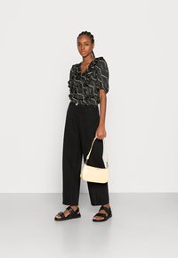 Blusa preta com babados e padrão branco, calças largas pretas e sandálias pretas. A segurar uma pequena mala amarela com acabamento brilhante.