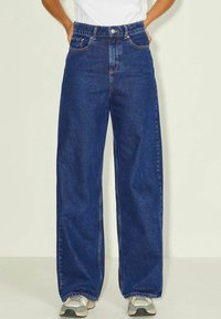 Oseba v temno modri širokih hlačah z sprednjimi žepi in sivih supergah na preprostem svetlem ozadju.