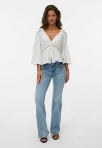 Donna che indossa una blusa bianca con scollo a V e dettagli in pizzo, jeans a campana blu chiaro e sandali neri con punta scoperta, in piedi su uno sfondo bianco.