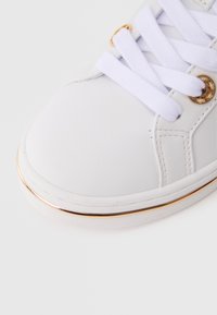 Sneaker bianca con tomaia sintetica liscia, lacci visibili, occhielli dorati e dettaglio dorato lungo la suola.
