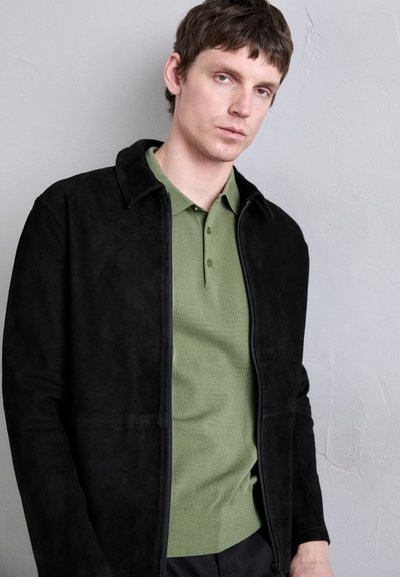 Veste en daim noir avec fermeture éclair à l'avant et col, portée sur un polo vert texturé. L'arrière-plan est gris clair.