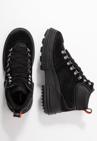 Botas de senderismo negras con parte superior de ante y suela de goma, con ojales plateados, cordones texturizados y una lengüeta con un acento naranja.