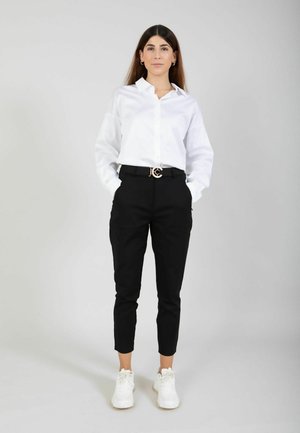 Mujer de pie con las manos en los bolsillos, vistiendo una camiseta blanca, pantalones negros, zapatillas blancas y un cinturón con una hebilla circular dorada sobre un fondo gris.