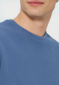 Κοντινό πλάνο ενός ατόμου που φοράει μπλε μπλούζα με στρογγυλή λαιμόκοψη και υφή, εμφανίζοντας το κάτω μέρος του προσώπου, τον λαιμό και το πάνω μέρος του στήθους.