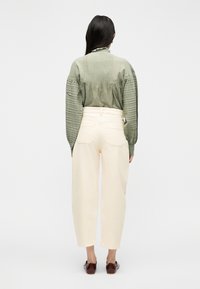 Γυναίκα με μακριά σκούρα μαλλιά που φοράει μια υφασμάτινη πράσινη μπλούζα, κρεμ παντελόνι ψηλόμεσο και σκούρα καφέ παπούτσια, γυρμένη με την πλάτη σε λευκό φόντο.