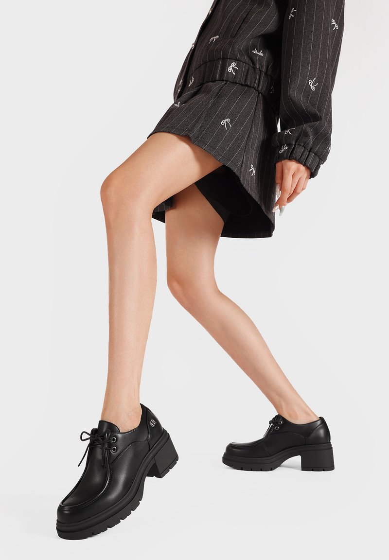 Scarpe in pelle nera con tacco spesso, punta rotonda e design con lacci. Suola in gomma con dettagli a tasselli. Indossate con una gonna a righe.