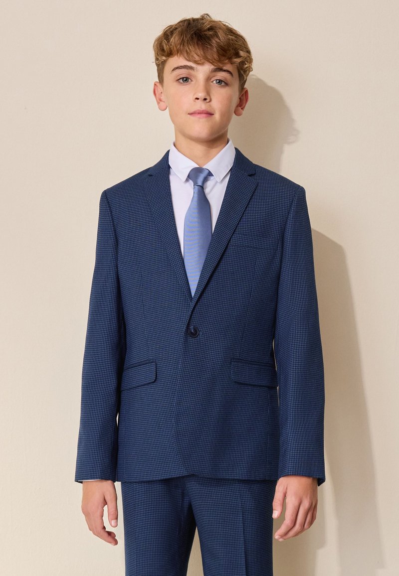 Niño vestido con un traje azul a cuadros y una corbata azul clara, de pie contra una pared beige, mirando hacia adelante.