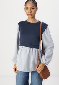 Mornarsko modra majica z črtasto tkanino na rokavih in peplum robom. Ima obsežne rokave in rjav čez ramo nahrbtnik.
