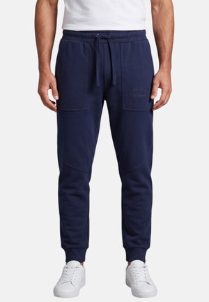 Hombre que lleva pantalones deportivos azul marino con bolsillos delanteros de parche y zapatillas blancas, de pie frente a un fondo liso.