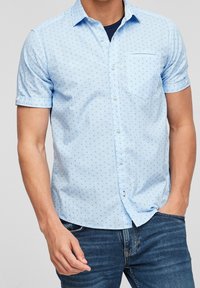 Camicia leggera a maniche corte di colore azzurro, realizzata in un tessuto leggero con un delicato motivo di piccole forme blu scuro, dotata di taschino anteriore e chiusura con bottoni.