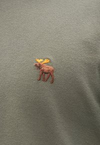 Olivovo zelená látka s malým vyšívaným losom v hnedej a žltej farbe, ktorá zobrazuje detaily stehov a farebného kontrastu.