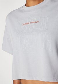 Skraćena siva majica od mekog materijala sa reljefnim natpisom "UNDER ARMOUR" u bijeloj i vibrantnoj crvenoj boji, okrugli izrez i kratki rukavi.