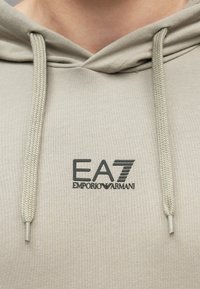 Közeli kép egy bézs kapucnis pulóverről, amelyen húzózsinórok vannak, és a mellkas közepén fekete EA7 Emporio Armani logó található.