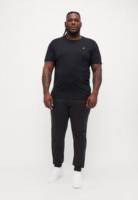 Schwarzes Baumwoll-T-Shirt mit Rundhalsausschnitt und kurzen Ärmeln, versehen mit einem kleinen goldenen Logo. Kombiniert mit schwarzen schmal geschnittenen Hosen und weißen Sneakern.