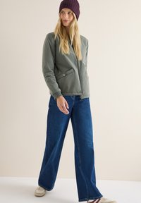 Olivengrøn bomberjakke med lynlås og trykknaplommer, parret med brede mørkeblå jeans og en mørk lilla strik-béret. Uformelt fodtøj synligt.