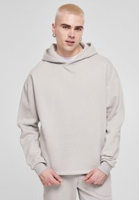 Lys grå hoodie lavet af blødt stof, med en stor hætte, løs pasform og ribbede manchetter. Modellen har matchende lys grå bukser på.