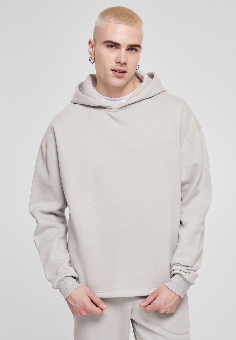 Lys grå hoodie lavet af blødt stof, med en stor hætte, løs pasform og ribbede manchetter. Modellen har matchende lys grå bukser på.
