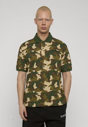 Jonge persoon met kort blond haar, gekleed in een groen-bruine camouflage polo en zwarte broek met logotekst.
