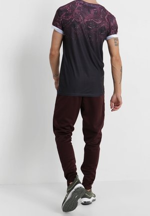 Man die een zwart en donkerrood grafisch T-shirt draagt, bordeauxrode joggers en groene sneakers, staand tegen een effen witte muur, gezien vanaf de achterkant.