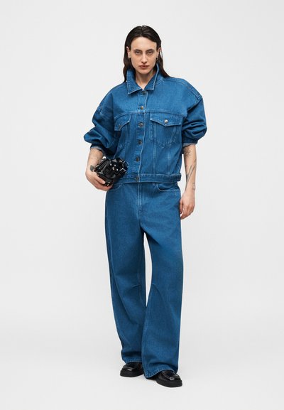 Άτομο που φοράει μπλε τζιν μπουφάν και τζιν παντελόνι φαρδιάς γραμμής, κρατώντας μια μαύρη τσάντα χειρός, στέκεται μπροστά σε ένα απλό λευκό φόντο.