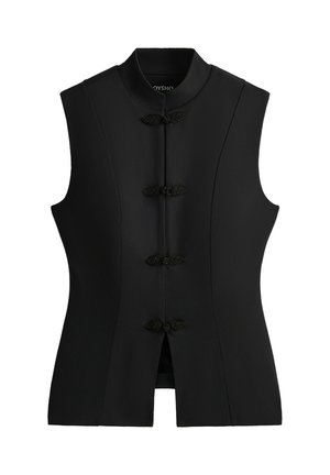 Femme debout portant un haut noir sans manches ajusté avec des boutons grenouille, des leggings capri noirs et des chaussures de sport noires sur un fond gris.